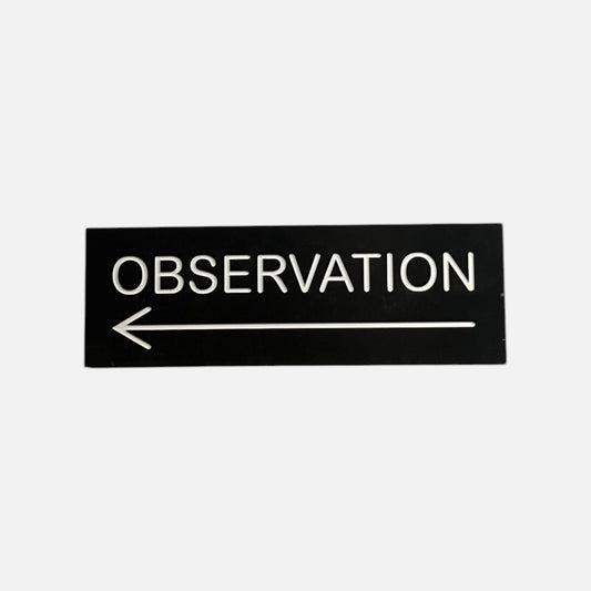 Long black OBSERVATION sign with raised white letters and a left pointing arrow beneath them, on a white background.
